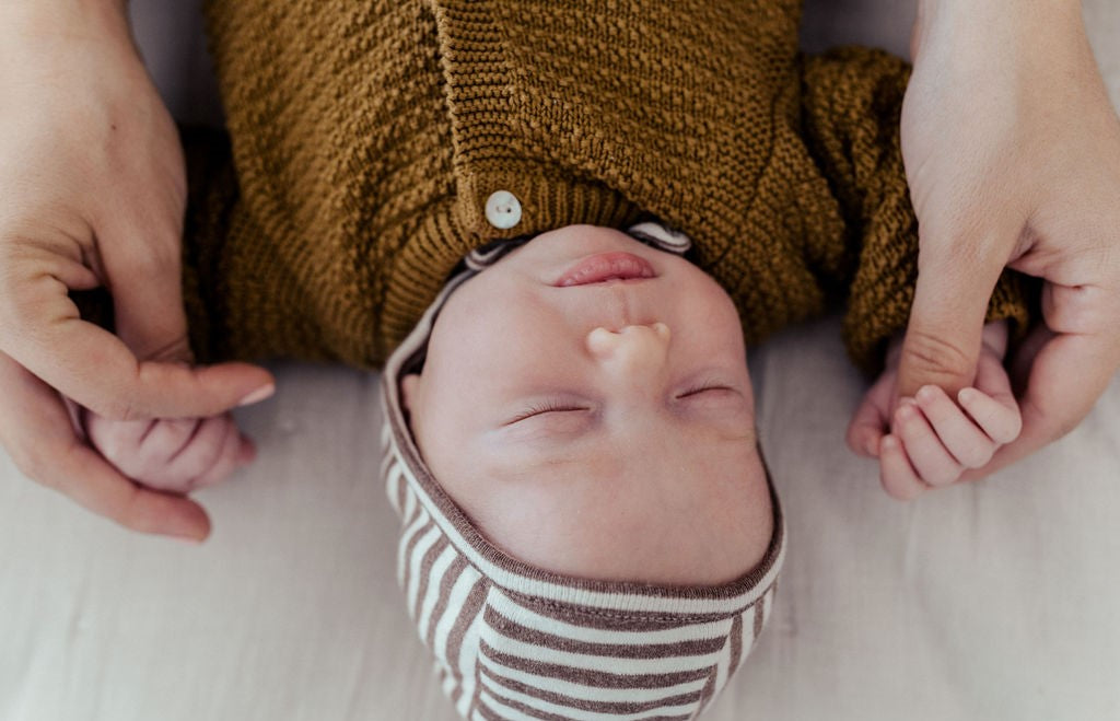 Photography close-up from above of a cute newborn baby with white skin sleeping wearing a striped dark brown and white hat from OiOioi and a brown cardigan from OiOiOi and holding hands of his mother 