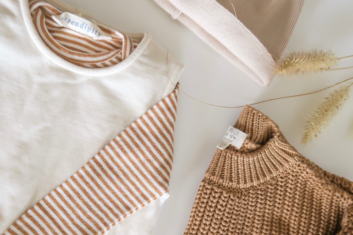 Photography from above of kid clothes from OiOiOi on a white background : on the left side a white kid hoodie from OiOiOi with brown  and white striped sleeves and on the right side a brown knitted folded kid sweater from OiOiOi and on the top a white and off-white baby hat and on the right side 2 stalks of wheat    