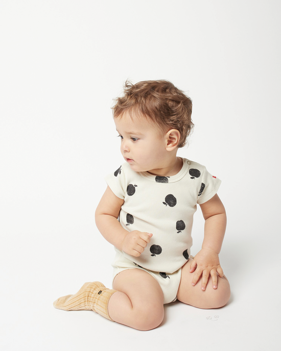 Baby with clear skin and brun hairs sitting and wearing a white body white black apple patterns from Bobo Choses on a white