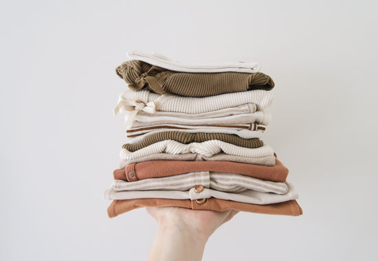 Hand with light skin color, holding folded organic cotton baby dresses in brownish and light natural colors from OiOiOi