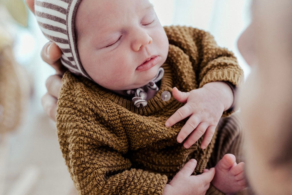 Baby with light skin tone, wearing brown cardigan from OiOiOi and a brown-white-striped hat.