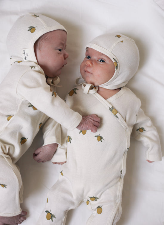 2 cute newborn babies laying on a white bed next to each other wearing both the same clothes, a white newborn hat with lemon patterns from Konges Sloejd and a white with lemon pattern long sleeve pyjama 