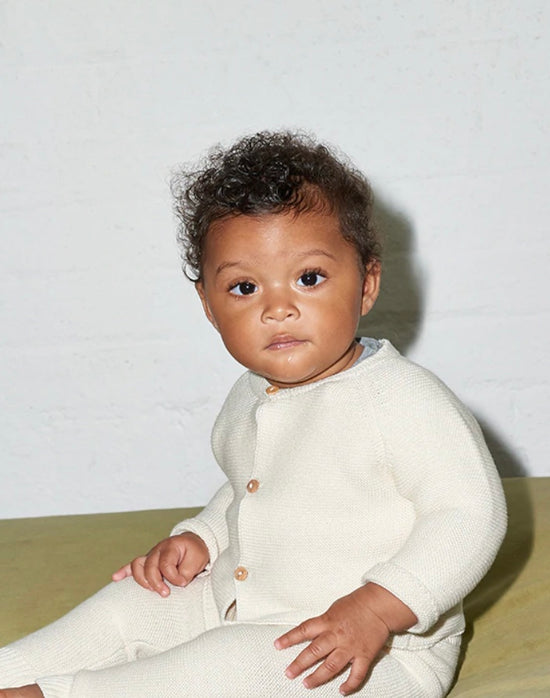 Baby with dark skin and dark curly hair sits on green underground, wearing white clothes from Vild house of little and OiOiOi