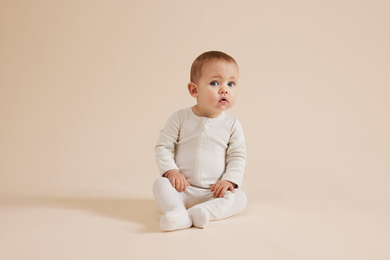 Baby with light skin color sits on floor with beige background and wears white children clothes from Wilson & Frenchy, OiOiOi