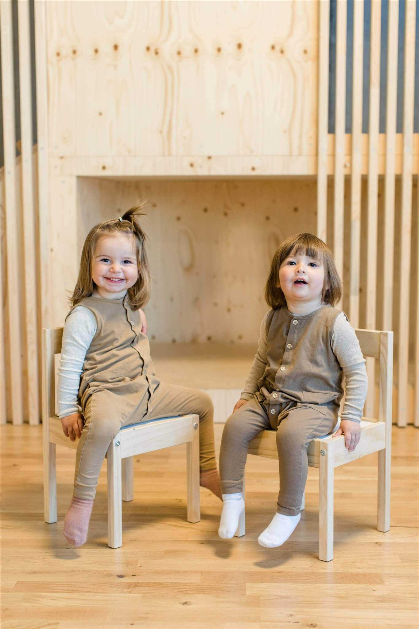 Two children with brown hair and brown clothes from OiOiOi each sitting smiling on wooden chairs